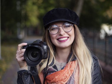 Bir refleks kamera sonbahar park ellerinde tutan makyaj ile gözlük ve şapkası giyen bir güzel şık neşeli genç sarışın fotoğrafçı portresi