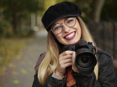Bir refleks kamera sonbahar park ellerinde tutan makyaj ile gözlük ve şapkası giyen bir güzel şık neşeli genç sarışın fotoğrafçı portresi