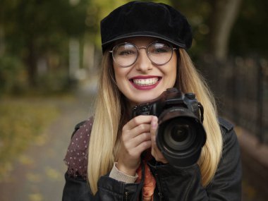 Portrait of a beautiful stylish cheerful young blonde photographer wearing glasses and a cap with makeup holding a reflex camera in her hands in autumn park clipart