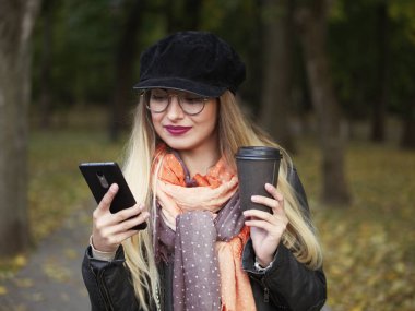 Sıcak bir içecek ve telefon görüşmeleri sonbahar soğuk parkta içen bir güzel şık genç sarışın bir deri ceketli yuvarlak gözlük makyaj ile portresi