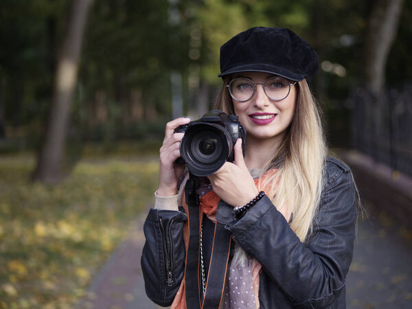 Portrait of a beautiful stylish cheerful young blonde photographer wearing glasses and a cap with makeup holding a reflex camera in her hands in autumn park