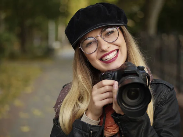 Bir refleks kamera sonbahar park ellerinde tutan makyaj ile gözlük ve şapkası giyen bir güzel şık neşeli genç sarışın fotoğrafçı portresi