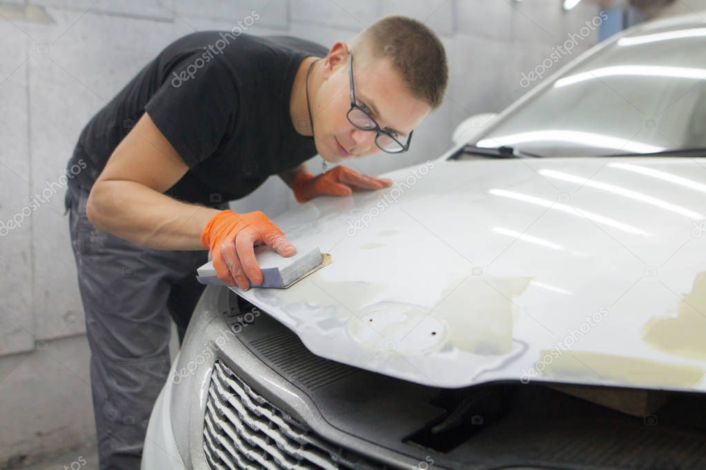 cute guy in black glasses with orange gloves displays the shape of the hood for the subsequent painting of the car. guy - a house painter working on a car in the garage 