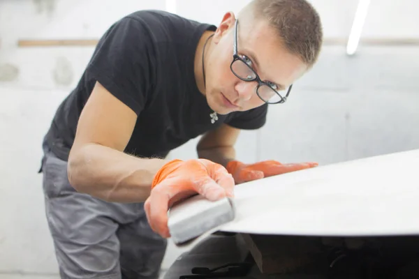 cute guy in black glasses with orange gloves displays the shape of the hood for the subsequent painting of the car. guy - a house painter working on a car in the garage