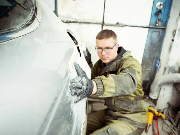 Cute guy in black glasses with black gloves displays the shape of the hood for the subsequent painting of the car. guy - a house painter working on a car in the garage