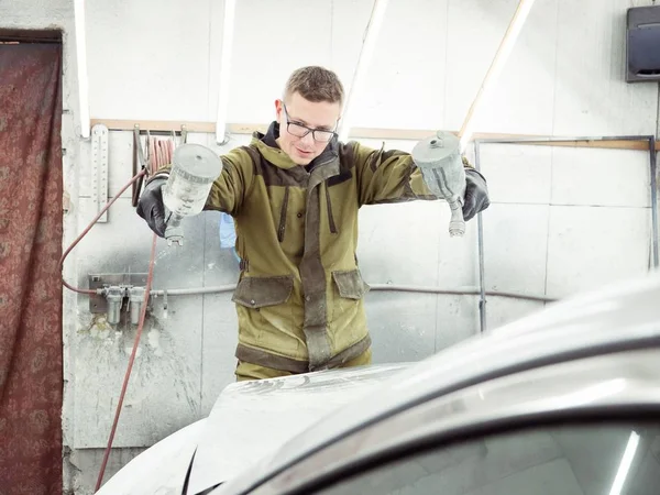 Cute guy in black glasses with black gloves displays the shape of the hood for the subsequent painting of the car. guy - a house painter working on a car in the garage