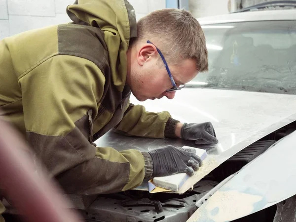 Cute guy in black glasses with black gloves displays the shape of the hood for the subsequent painting of the car. guy - a house painter working on a car in the garage