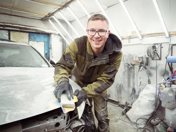 Cute guy in black glasses with black gloves displays the shape of the hood for the subsequent painting of the car. guy - a house painter working on a car in the garage