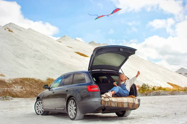 Kız arabada otururken dağlarda uçurtma fırlatıyor.