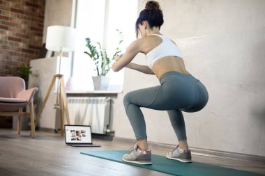 Genç, spor düşkünü kadının internetteki spor eğitmeni modern laptop ekranı var. Sağlıklı yaşam tarzı konsepti, online fitness ve spor dersleri. Çömelmeler, sallanan kalçalar