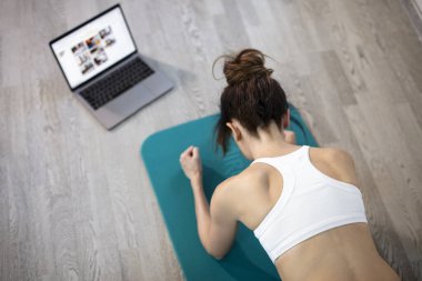 Evde eğitim gören zayıf genç bir kadın. İnternette dizüstü bilgisayarla çalışıyor. Ve spor paspası. Çevrimiçi eğitim, çevrimiçi fitness, evde kalmak, karantina, online eğitim. Plank egzersizi.