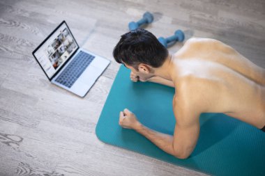 Evde genç sıska adam eğitimi. Bilgisayarlı bir hasırın üzerinde egzersiz yapıyor. Tahta egzersizi yapıyor. Güçlü eller. Çevrimiçi eğitim, çevrimiçi fitness, evde kalmak, karantina, online eğitim. Plank egzersizi.