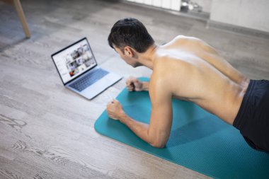 Evde genç sıska adam eğitimi. Bilgisayarlı bir hasırın üzerinde egzersiz yapıyor. Tahta egzersizi yapıyor. Güçlü eller. Çevrimiçi eğitim, çevrimiçi fitness, evde kalmak, karantina, online eğitim. Plank egzersizi.