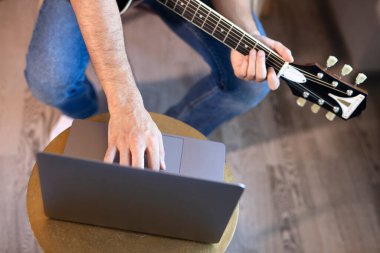 Genç bir adam koltukta bir odada oturur, online gitar çalmayı öğrenir. Çevrimiçi eğitim, çevrimiçi müzik dersleri, online vokal ve gitar eğitimi. Evde kal..