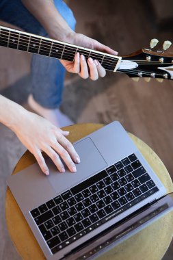 Genç bir kadın koltukta bir odada oturuyor ve internetten gitar çalmayı öğreniyor. Çevrimiçi eğitim, çevrimiçi müzik dersleri, online vokal ve gitar eğitimi. Evde kal..