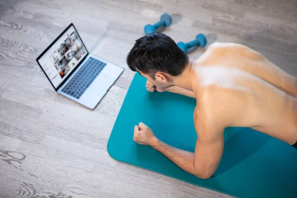 Evde genç sıska adam eğitimi. Bilgisayarlı bir hasırın üzerinde egzersiz yapıyor. Tahta egzersizi yapıyor. Güçlü eller. Çevrimiçi eğitim, çevrimiçi fitness, evde kalmak, karantina, online eğitim. Plank egzersizi.
