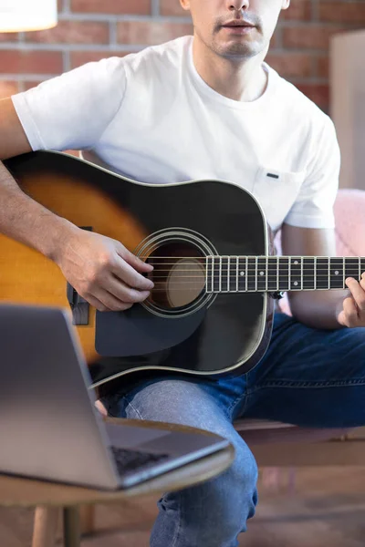 Young man sits in a room on an armchair, learns to play guitar online. Online education, online music lessons, online vocal and guitar training. Stay home.