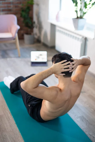 Young slim man training at home. Workout online fitness lessons on a mat with a laptop, doing russian twisting and sit-ups. Online training, online fitness, stay home, quarantine, online training.