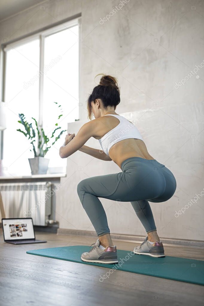 Young sporty slim woman has internet video online fitness training instructor modern laptop screen. Healthy lifestyle concept, online fitness and sport lessons. Squats, swing buttocks