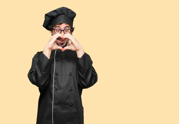 young crazy man as a chef smiling, looking happy and in love, making the shape of a heart with hands. Front view.