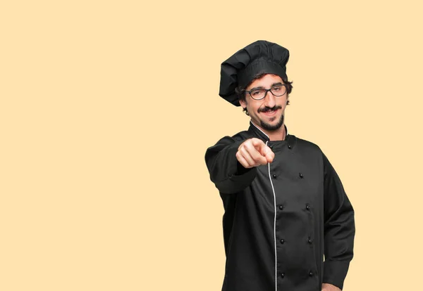 young crazy man as a chef smiling happily and pointing forward, choosing you.