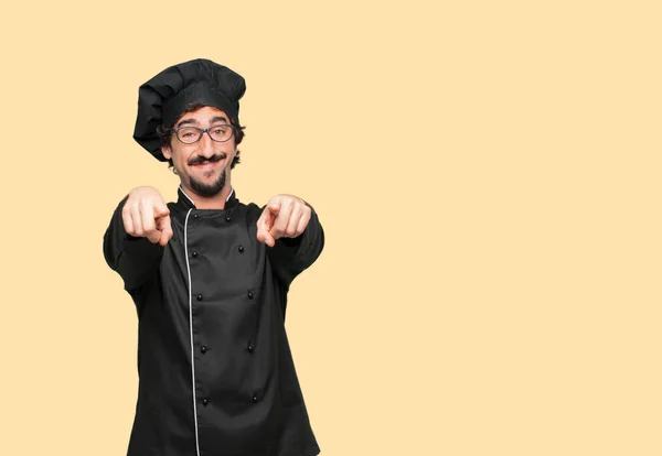 young crazy man as a chef smiling happily and pointing forward with both hands, choosing you.