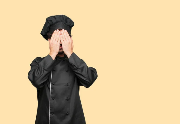 young crazy man as a chef with a serious, scared, frightened expression, covering eyes with both hands.