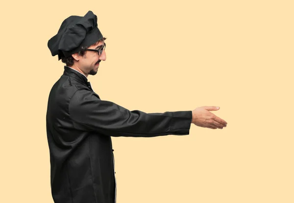 young crazy man as a chef with a smiling, confident, proud, satisfied and friendly expression offering a handshake, closing a good deal, or greeting and welcoming you. Lateral or side view.