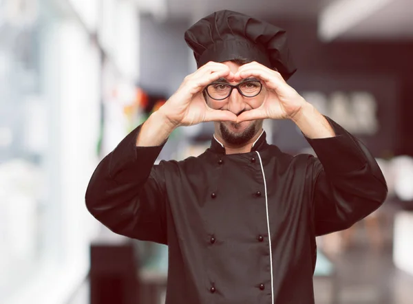 young crazy man as a chef standing sideways, smiling, looking happy and in love, making the shape of a heart with hands. Side or lateral view.