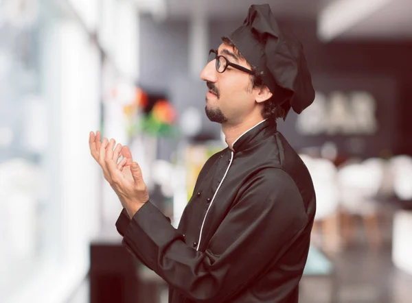 young crazy man as a chef looking stressed and frustrated, looking upwards and holding both hands open in front expressing dismay and disbelief.