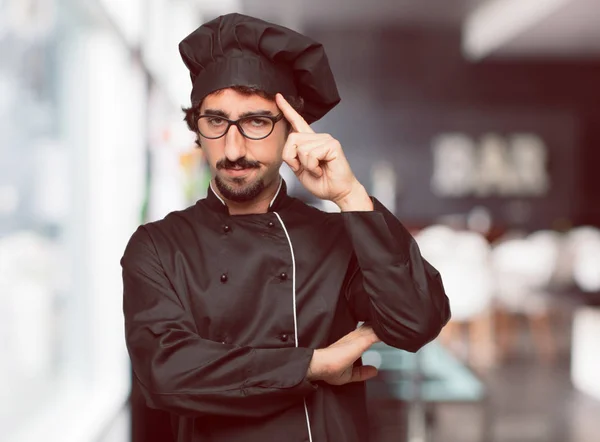 young crazy man as a chef Concentrating hard on an idea, with a serious far-off look, thinking with arm crossed in front and index finger pointing to forehead.