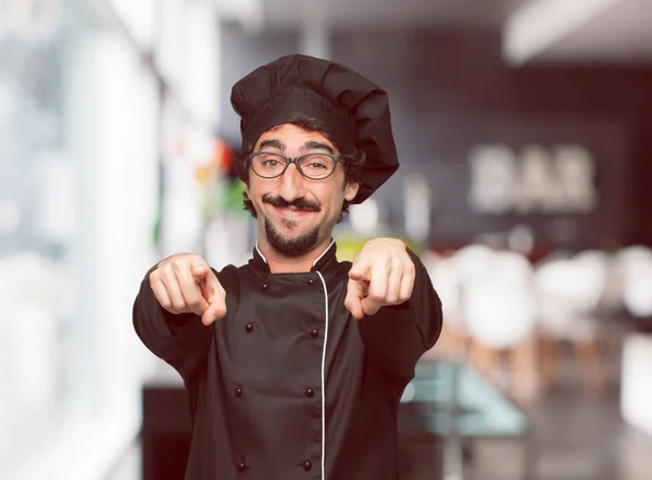young crazy man as a chef smiling happily and pointing forward with both hands, choosing you.
