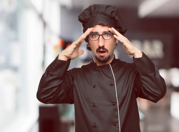 young crazy man as a chef with a surprised, amazed expression, holding both hands to forehead and mouth wide open in shock.