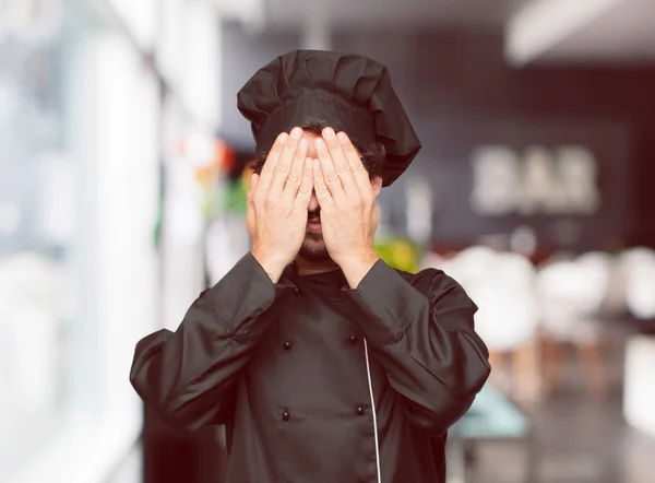 young crazy man as a chef with a serious, scared, frightened expression, covering eyes with both hands.