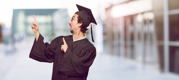 young graduated man smiling and pointing to the side with both hands, towards the place where the publicist may show a concept.