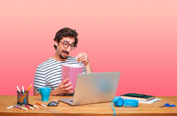 young crazy graphic designer on a desk with a laptop and with a pop corn bucket