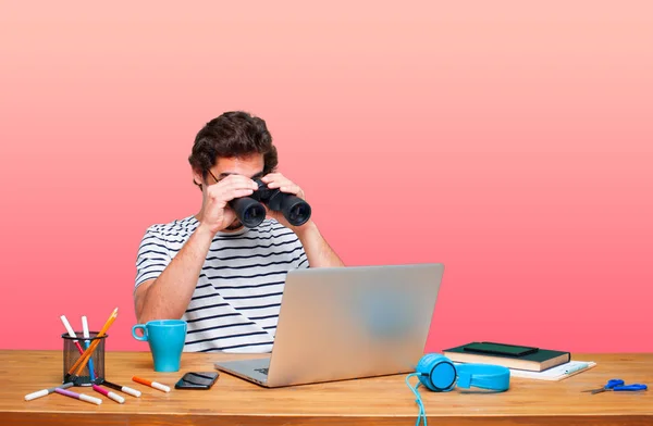 young crazy graphic designer on a desk with a laptop and with binoculars