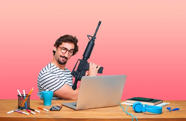 young crazy graphic designer on a desk with a laptop and with a rifle. Conflict concept.