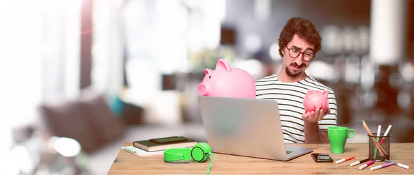 young crazy graphic designer on a desk with a laptop and with a piggy banks