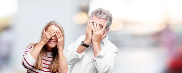 senior cool husband and wife with a scared, frightened expression, covering eyes with both hands, peeking.