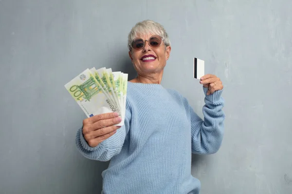 senior cool woman with a credit card against grunge cement wall. money or savings concept.