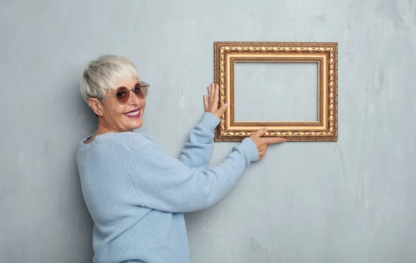 senior cool woman with a baroque frame against grunge cement wall.