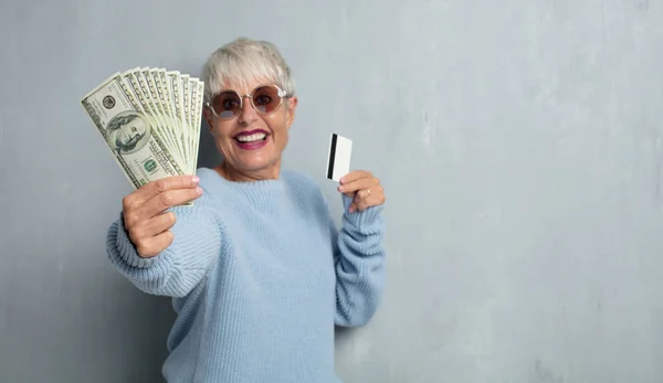 senior cool woman with a credit card against grunge cement wall. money or savings concept.