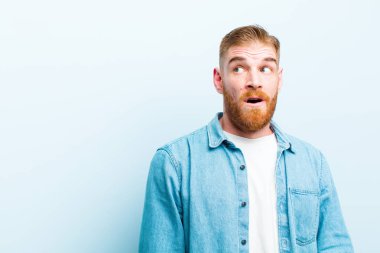 genç kırmızı baş adam şok duygu, mutlu, şaşırmış ve şaşırttı, yumuşak mavi arka plan karşı açık ağız ile yan bakıyor