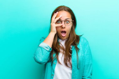 genç kadın şok görünümlü, korkmuş ya da korkmuş, el ile yüz kapsayan ve mavi arka plan karşı parmaklar arasında gözetleme