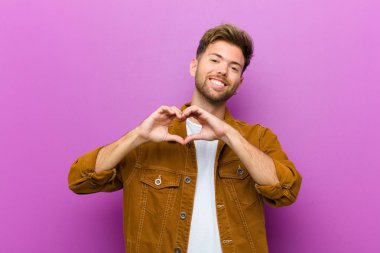 young man smiling and feeling happy, cute, romantic and in love, making heart shape with both hands against purple background