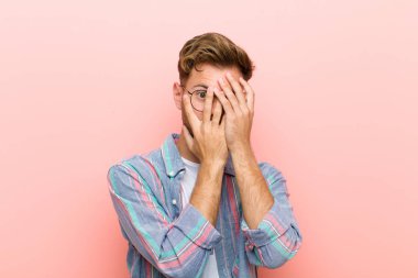 young man feeling scared or embarrassed, peeking or spying with eyes half-covered with hands against pink background