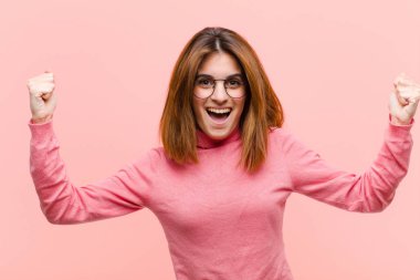 young pretty woman shouting aggressively with an angry expression or with fists clenched celebrating success against pink background