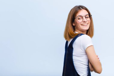 Genç, güzel bir kadın neşeyle gülümsüyor, mutlu, tatmin olmuş ve rahatlamış hissediyor, kollarını kavuşturup mavi arka plana bakıyor.