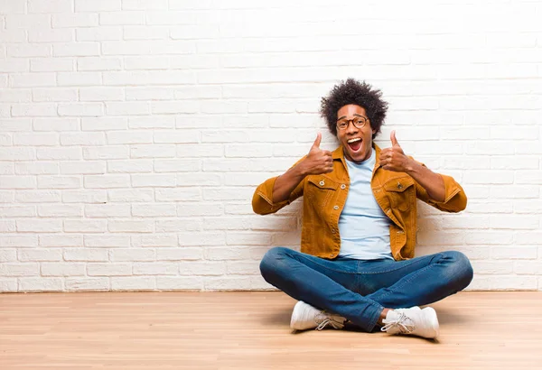 若い黒人男性は 自宅で床に座って 両方の親指で 肯定的 自信と成功を見て広く微笑んでいます — ストック写真